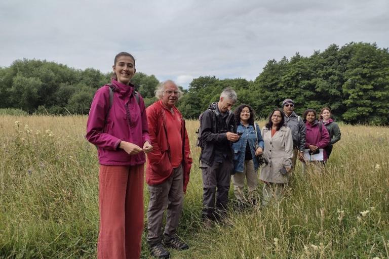 A group of eight people stand together in a grassy field surrounded by tall wildflowers and green trees under a cloudy sky. The individuals are dressed in outdoor clothing, including jackets and backpacks, and appear to be engaged in an outdoor activity or nature walk.