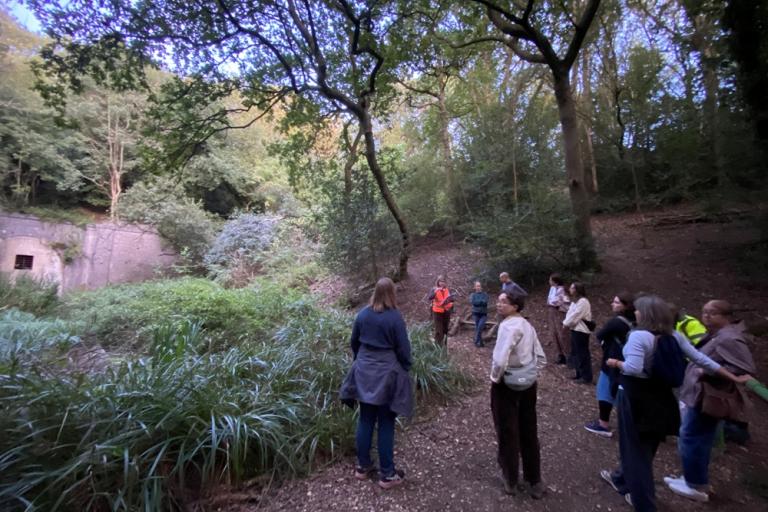 A group of people stand on a woodland path surrounded by mature trees and dense vegetation, listening to a guide in a bright orange high-visibility vest. 