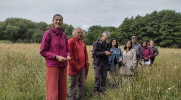 A group of eight people stand together in a grassy field surrounded by tall wildflowers and green trees under a cloudy sky. The individuals are dressed in outdoor clothing, including jackets and backpacks, and appear to be engaged in an outdoor activity or nature walk.